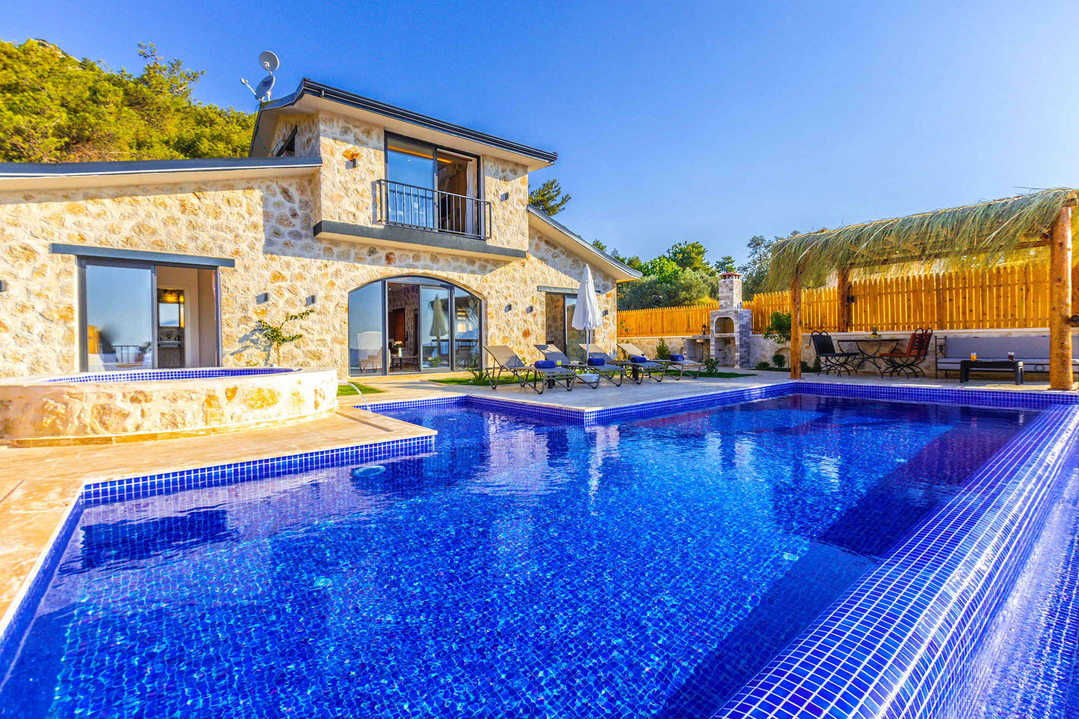 Gran piscina azul, con desbordamiento, tan grande como la fachada de una casa tipo Chalet, de dos plantas; con jacuzy, barbacoa, pérgola.